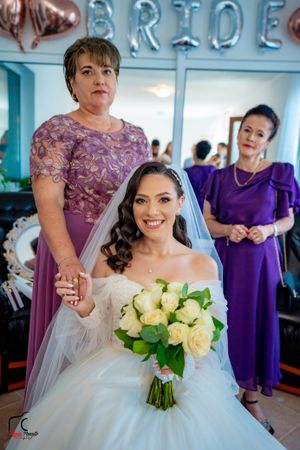 Bride holding cream roses poses with bridesmaids in purple dresses during wedding preparations.