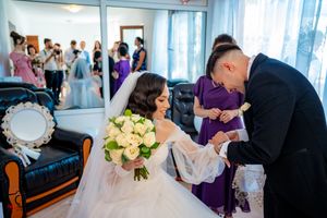 Wedding ceremony scene with attendees in formal attire gathered in a sunlit room.