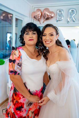 Bride in white gown poses with guest wearing vibrant floral dress in bridal suite.