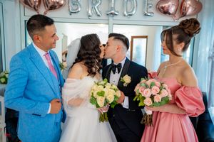 Wedding party members pose in a room decorated with 'BRIDE' balloons, featuring pink dresses and a light blue suit.