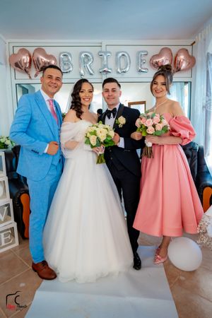 Wedding celebration scene with bridal party wearing pink dresses and light blue suit against white wall decor.