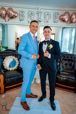 Two people in formal attire pose in a room with black leather furniture and balloon decorations.