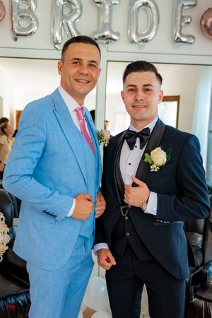 Two people in contrasting blue and black suits pose under silver balloon letters spelling 'BRIDE'.