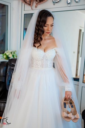 A bride in an off-shoulder wedding gown holds nude-colored heels while wearing a flowing veil.