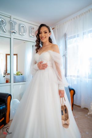 Bride in princess-style wedding dress holds nude heels in front of silver BRIDE balloon letters.