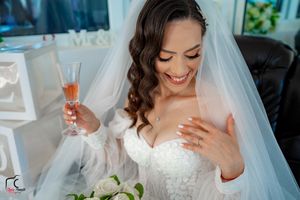 Bride in white lace dress smiles while holding champagne and touching her heart.