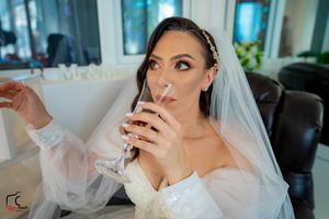 Bride in white dress and veil sips champagne while looking up thoughtfully.