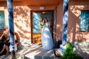 Bride stands in white gown holding bouquet on porch of terracotta-colored building.