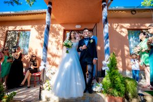 A wedding celebration scene outside a terracotta colored building with blue sky and greenery visible.
