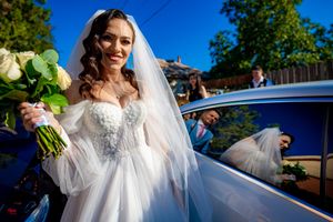 Bride in white lace dress posing next to a blue car outdoors.