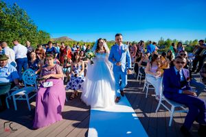 Outdoor wedding ceremony with attendees in blue and purple attire gathered on a wooden deck.