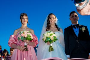 Wedding party members holding floral bouquets during outdoor ceremony against blue sky.