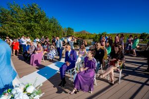 Panoramic view of outdoor wedding ceremony with guests gathered on paved area surrounded by trees.