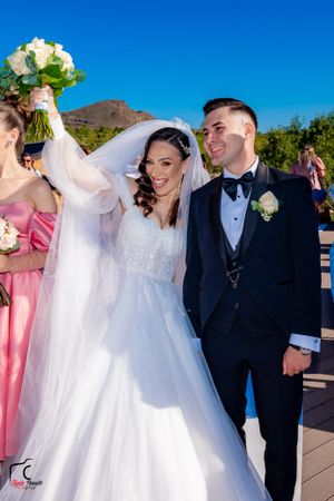 Newlyweds celebrate joyfully outdoors with white floral bouquet against mountain landscape.