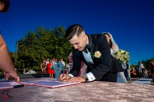 Groom in black tuxedo signs marriage certificate during outdoor ceremony.