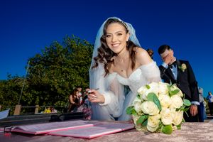 Bride with bouquet signing marriage certificate under bright blue sky.
