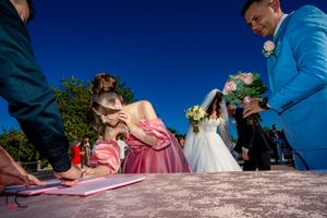 Wedding party members gather around to sign marriage documents in natural lighting.