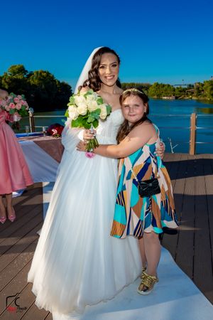 Wedding party members gather on waterfront deck with white dress and colorful attire.