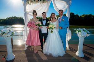 Outdoor waterfront wedding ceremony with white fabric arch and floral decorations at sunset.