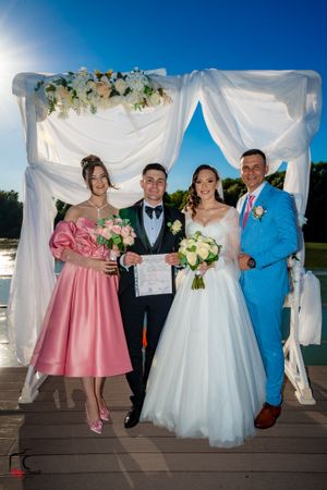 Wedding group photo on wooden deck with white and pink formal attire against blue sky.