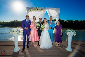 Wedding party members in formal attire pose on waterfront deck with decorative arch.