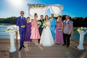 Wedding celebration group photo with mixed formal attire on lakeside wooden deck.