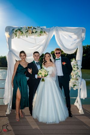 Wedding party poses for photos on a lakeside deck with white draped arch and floral decorations at sunset.