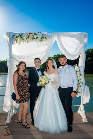 Wedding group gathers under white fabric arch with floral accents on wooden deck by water.