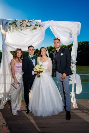 Outdoor wedding party photos on wooden deck with white fabric arch and blue sky backdrop.