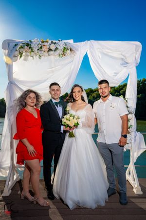 Wedding celebration group shot on waterfront deck with white ceremony arch and summer sky.