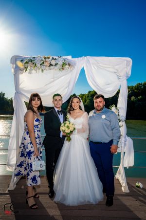 Wedding party members pose together under decorated white arch on lakeside deck.