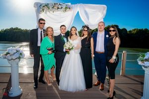 Wedding party gathering on outdoor deck with decorated arch and waterfront scenery.