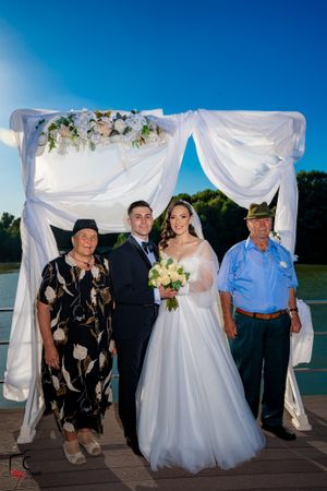 Wedding party poses on a waterfront deck with white draped arch and floral decorations at sunset.