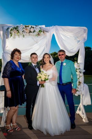 Wedding group portraits taken on wooden deck with white fabric arch and floral arrangements.