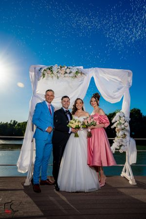 Outdoor waterfront wedding party posing under white fabric arch with pink and white floral arrangements.