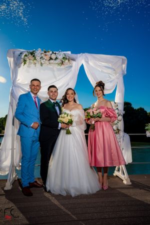 Wedding party gathered under white fabric arch with lake backdrop during sunset ceremony.