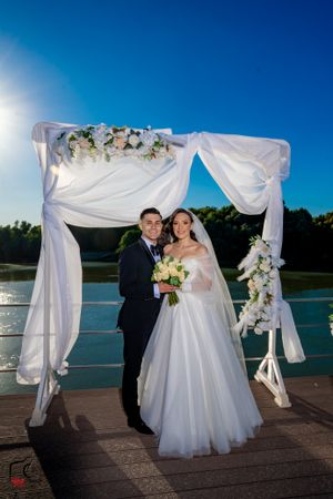Elegant lakeside wedding ceremony with white draped arch and floral arrangements against blue sky.