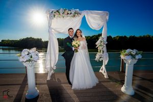 Waterfront wedding setup with white columns, flowing fabric, and floral arrangements on wooden dock.
