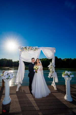 Sunlit wedding ceremony on lakeside dock with white draped arch and decorative columns.