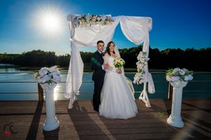 Sunset wedding ceremony overlooking lake with white fabric arch and floral decorations.