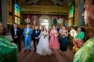 Wedding group photo in a traditional Orthodox church with colorful religious iconography.
