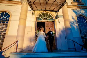 Wedding party gathers on the steps of a cream-colored church with arched windows and ornate architecture.
