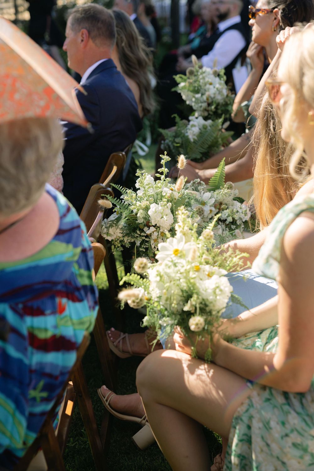 Wedding guests in outdoor seating hold beautiful bouquets of white and green flowers during a summer ceremony.