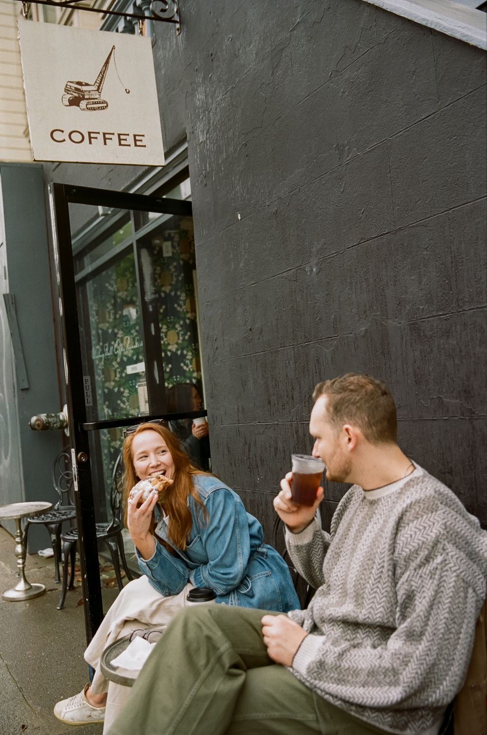 Two people enjoy coffee and pastries while sitting outside a coffee shop with a black wall and minimalist signage.