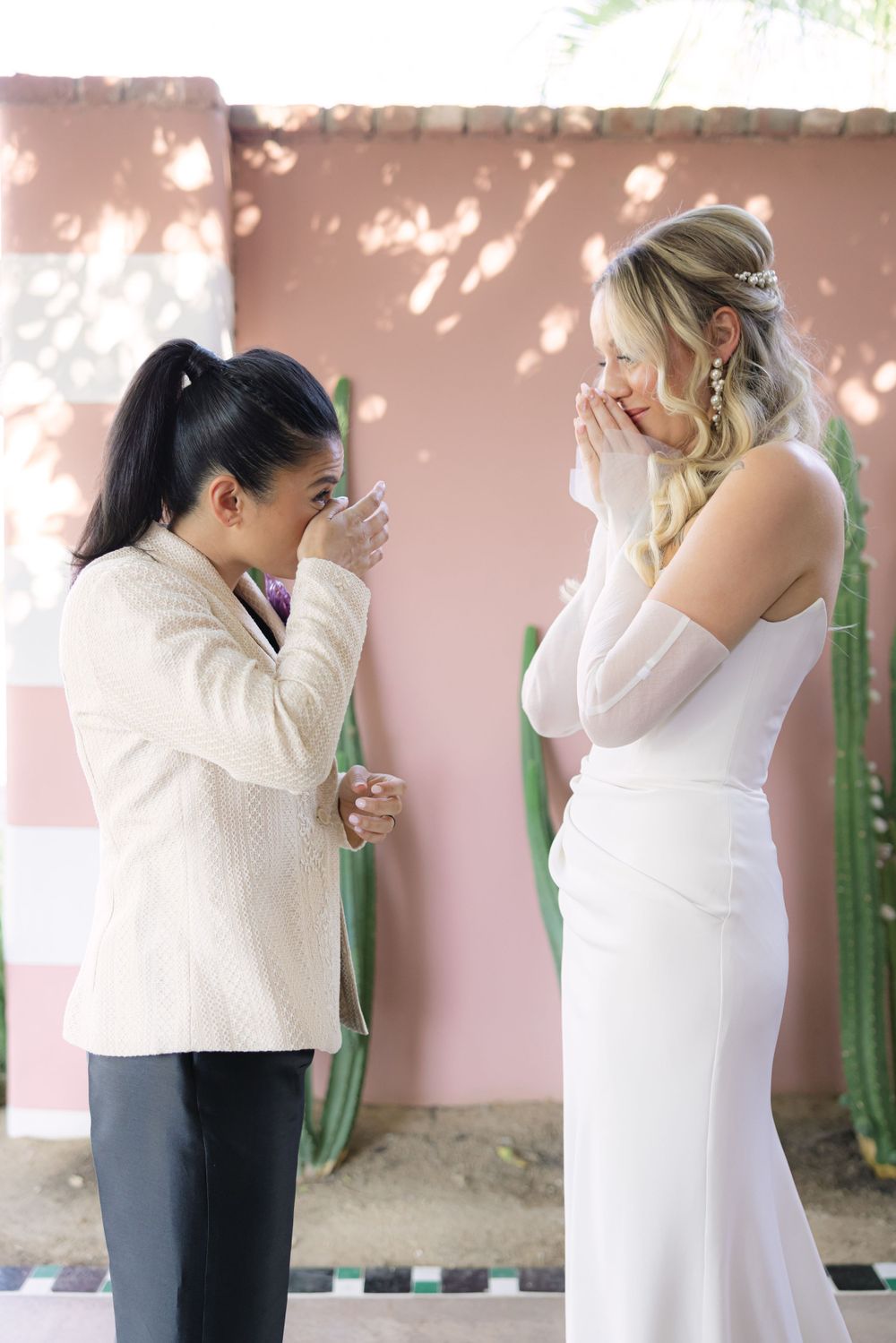 Two people share an emotional moment outdoors, one wearing an elegant white gown and the other in a cream colored jacket.
