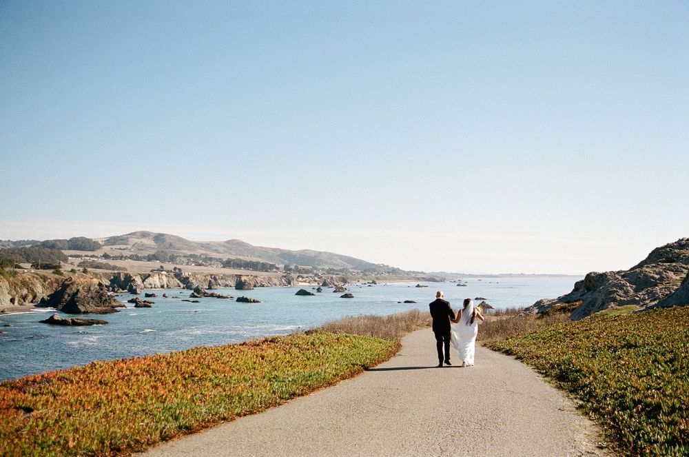 Two silhouettes walk along a coastal path bordered by orange wildflowers with boats anchored in the harbor beyond.