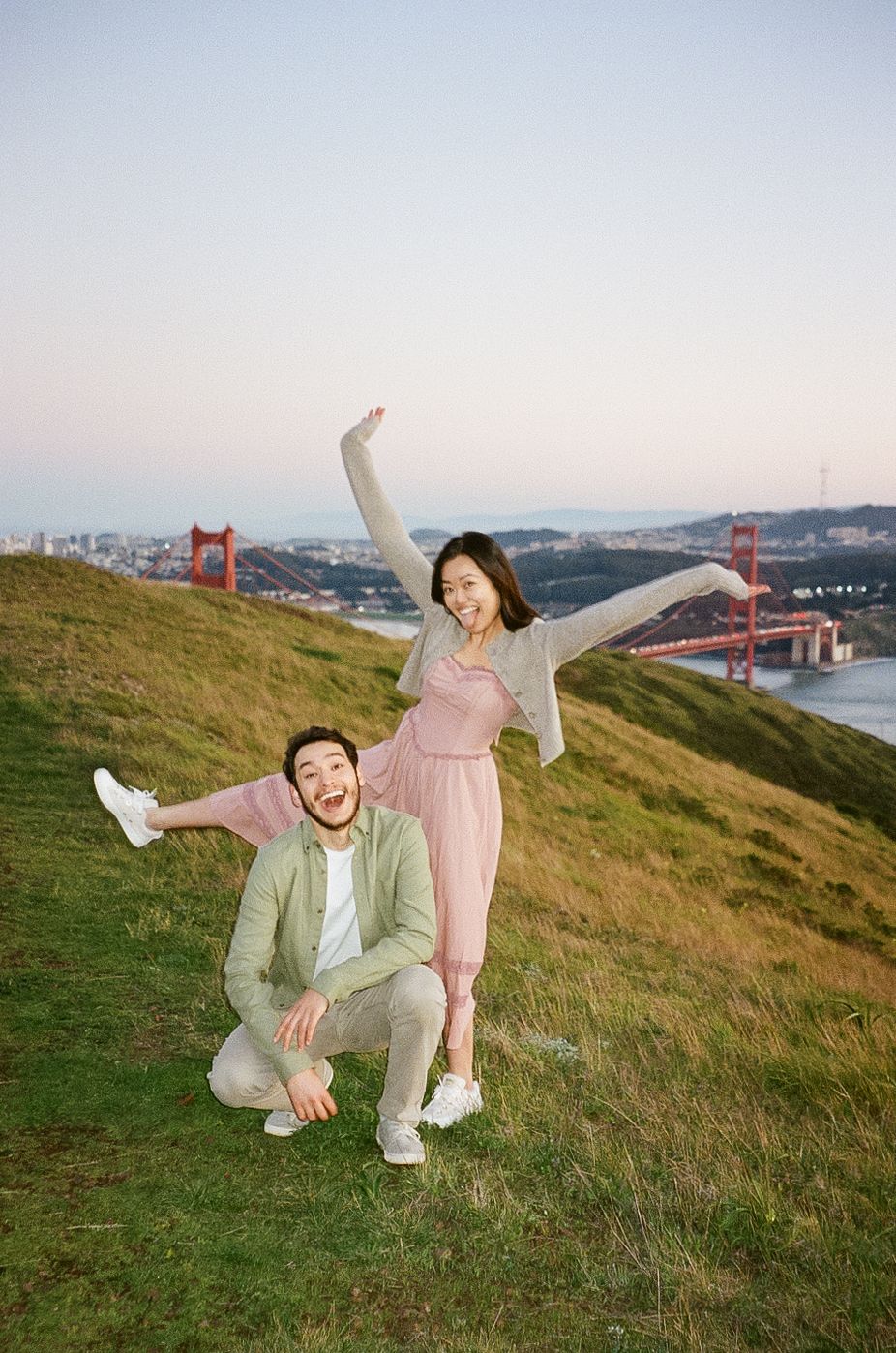 A couple poses playfully on a grassy hill overlooking the Golden Gate Bridge in San Francisco during sunset.