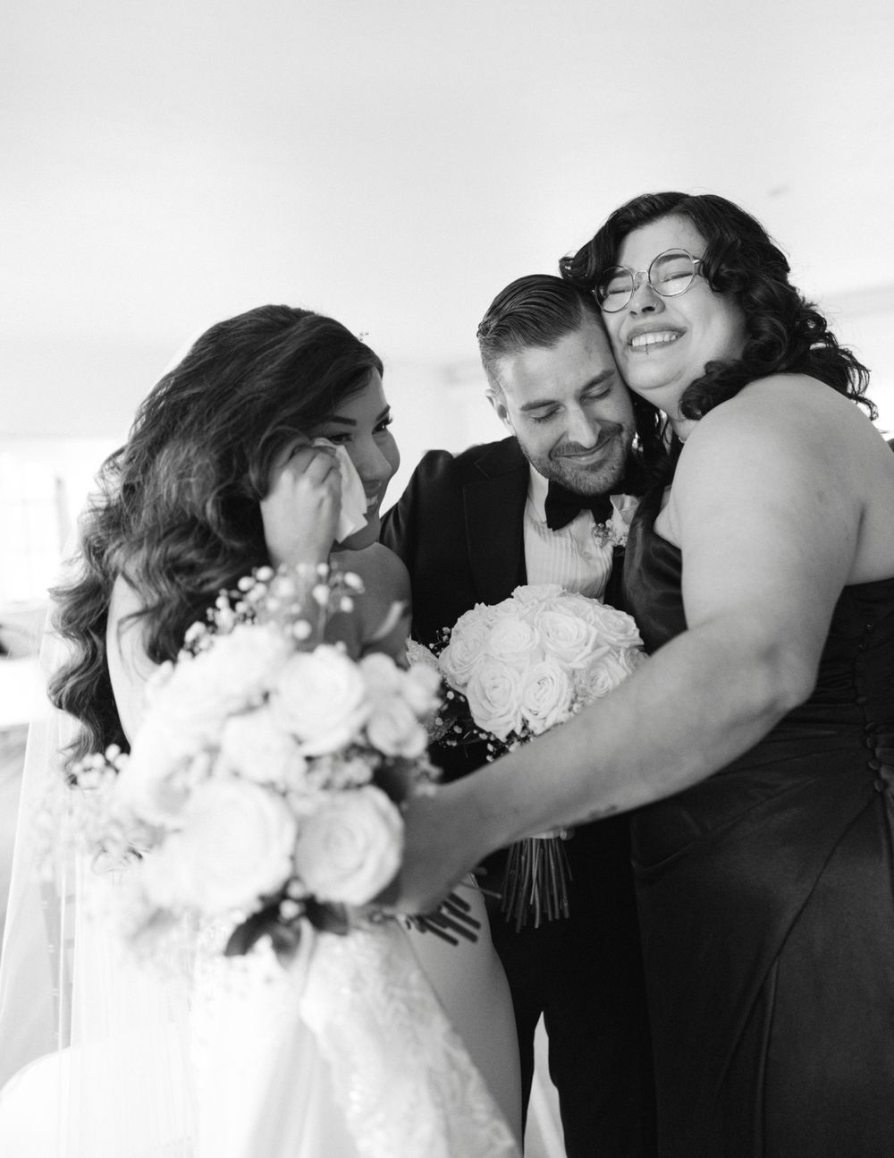 Black and white wedding photo showing joyful moment with wedding party sharing laughter while holding a bouquet of flowers.