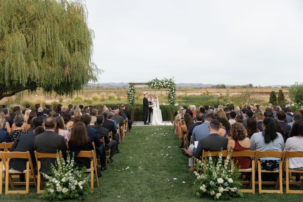 A scenic outdoor wedding ceremony with wooden chairs and floral arrangements under a weeping willow tree in a garden setting.