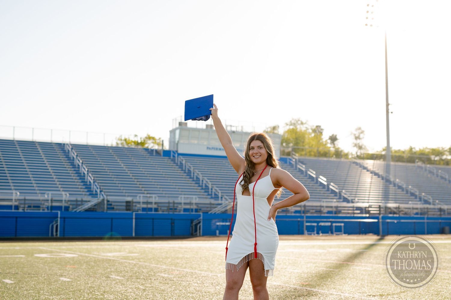Graduate raises blue cap in celebration on football field with stadium seating behind.